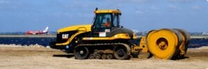 A yellow and black construction vehicle is parked on a sandy surface near a body of water. In the background, a red and white airplane can be seen on a runway. The sky is cloudy with a hint of blue.