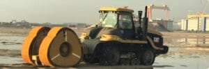 A road compactor machine is parked on a construction site. It has large yellow rollers in the front and visible CAT branding. In the background, there's industrial equipment and a building under a clear sky.