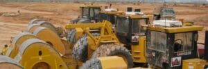 A row of large yellow construction vehicles, including rollers and bulldozers, parked on a dirt construction site, with cloudy skies in the background and additional machinery visible in the distance.