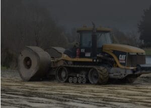 A large Caterpillar tractor is parked on a muddy construction site. The machine has large spiked metal wheels and visible tracks, with a dark cloudy sky and trees in the background.