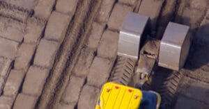 Aerial view of a yellow construction vehicle with two large metallic rollers compacting a surface with square stones, leaving parallel tracks.