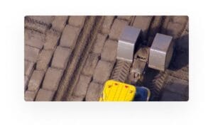 Aerial view of a yellow construction vehicle with two large cylindrical rollers, compacting a dirt surface with rectangular patterns.