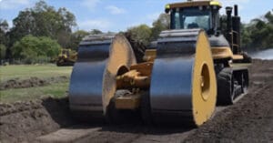 A large construction vehicle with wide, cylindrical rollers is compacting soil on a dirt road in a park-like area, surrounded by greenery and trees under a clear blue sky.