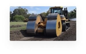 A large construction vehicle compacts soil on a site. It has twin metal rollers and is operating on a dirt path, surrounded by greenery and trees in the background, under a partly cloudy sky.