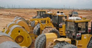 Heavy construction machinery, including yellow steamrollers and graders, are seen on a vast dirt construction site under a cloudy sky. The equipment is positioned in a row, indicating active land development or roadwork.