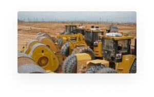 A row of yellow construction vehicles with large wheels are parked on a dirt terrain, with an expansive landscape in the background under a cloudy sky.