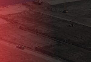 Aerial view of a construction site with large earth-moving equipment working on a vast expanse of dark soil. Red tint overlays the image, creating a dramatic effect. Sparse structures and machinery are visible in the distance.