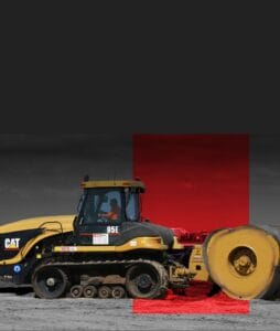 A construction vehicle operates on a sandy terrain. The image has a black and red overlay, highlighting the earth compactor. The sky is overcast, creating a dramatic backdrop for the machinery.