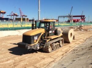 A yellow CAT construction vehicle with tracks and a large roller attachment is smoothing the sandy ground at a construction site. In the background, there are cranes, a green fence, and blue sky.