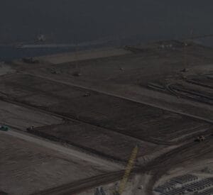 Aerial view of a large construction site near a body of water. The site features multiple cranes and heavy machinery working on leveled dirt and sand areas, with sections marked out for development. The water borders the upper side of the image.