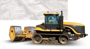 A heavy-duty, yellow track-type tractor with a black cab pulling a large soil compactor on a white background. The compactor is equipped with two massive metal rollers. The image suggests construction or land development.