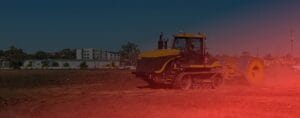 A yellow construction vehicle with caterpillar tracks operates on a dirt field, with a roller attached at the rear. Buildings and trees are visible in the background under a clear blue sky, and a red light effect covers part of the image.