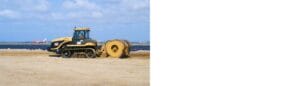 A construction vehicle with tank-like treads is rolling a large cylindrical object on a sandy construction site near the water. In the background, a red and white airplane approaches a runway beneath a partly cloudy sky.