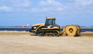 A construction vehicle is working on a sandy area near the water, with a large roller attached. In the background, a red airplane is taking off or landing. The sky is partly cloudy.