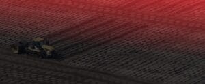 Aerial view of a tractor plowing a large, patterned field, illuminated with a reddish hue from the sunset. The repetitive texture of the soil creates a striking visual effect.