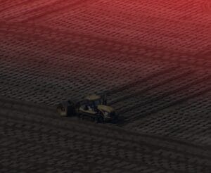 Aerial view of an agricultural field with a tractor and machinery working on patterned soil. The upper portion of the image is tinged with a red hue, adding a dramatic effect to the rural landscape.