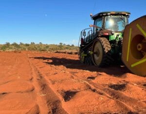 A green tractor with large wheels works on a vast, reddish-brown dirt field under a clear blue sky. Vegetation and trees are visible in the distance.