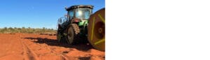 A green tractor with large wheels moves across a red dirt field under a clear blue sky, showcasing the practical applications of modern farming machinery. The landscape remains flat, with sparse vegetation fading into the distance.