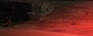 A large open pit mine with earth-moving machinery is visible in a barren, rocky landscape. The image has a red overlay, giving it a dramatic effect, with a digger and a truck near piles of dirt and stones.