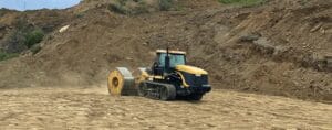 A yellow construction vehicle with tracks is compacting the ground at a site. It has a large steel roller attached at the rear. The background shows a dirt slope and blue sky.