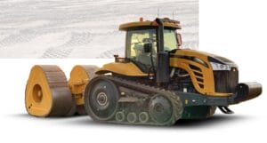 A large, yellow tracked tractor towing a heavy roller compactor on a construction site. The machinery is designed for flattening and preparing surfaces, with visible tracks and a robust frame. The background shows faint tire tracks on dirt.