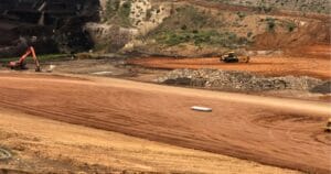 Construction site with excavators and bulldozers working on a large area of cleared land. Earth and gravel piles are visible. The terrain is uneven, with fine red dirt covering much of the ground, surrounded by a barren landscape.