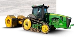 A green and yellow agricultural tractor with caterpillar tracks is pulling a large yellow compactor on a sandy surface. The machinery is designed for heavy-duty farming or construction work.
