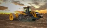 A large yellow tractor with caterpillar tracks stands on a muddy field at sunset. The sky is filled with clouds, and the warm light casts a glow on the tractor's surface.