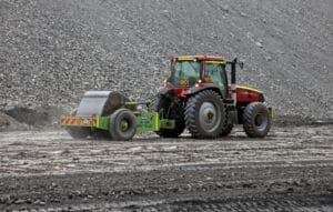 A red tractor pulling a roller compactor on a gravel surface. The area is surrounded by heaps of gray rocks, indicating a construction or roadwork site under overcast skies. Dust is visible behind the roller.