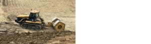 A construction vehicle with caterpillar tracks and a large drum roller is smoothing a dirt surface on a construction site. The ground is uneven, indicating ongoing land preparation work.
