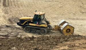 A construction vehicle with large wheels and a yellow body is moving across a dirt terrain. The ground is uneven and appears to be under construction or being leveled. There is a fire extinguisher attached to the side of the vehicle.