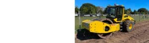 A yellow road roller is parked on a grassy construction site. It has large cylindrical wheels and is next to a fence, surrounded by trees and a clear blue sky in the background.