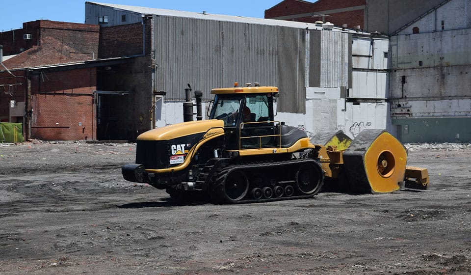 A yellow and black Caterpillar construction vehicle with tracks pulls a large, circular weight across a dirt lot in an industrial sector, framed by brick and metal buildings. The scene exudes business efficiency amidst the rugged charm of heavy machinery.