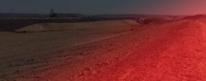 A vast, barren landscape with reddish soil under a hazy sky. Earth-moving equipment is barely visible in the distance, shaping the rugged terrain for a sustainable energy future. A red light effect partially overlays the right side of the image.