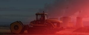 A construction vehicle with a large roller attachment operates near a power plant, where smoke billows from its cooling towers. Cast in an eerie red light, the scene underscores the importance of safeguarding environmental health amidst industrial operations.