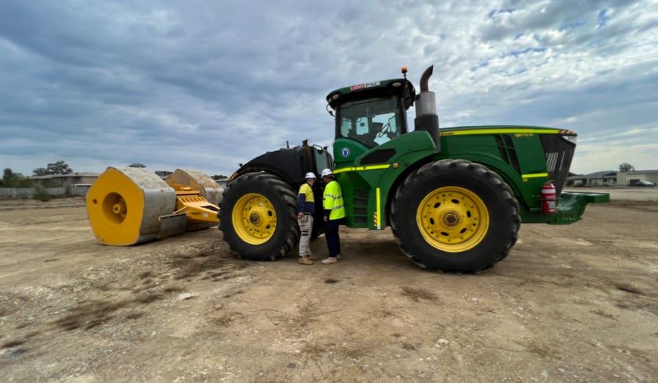 A large green and yellow tractor with construction equipment is parked on a dirt site, harnessing the potential of HEIC technology. Two people in safety gear and helmets stand beside the tractor under a cloudy sky, ensuring environmental health standards are met.