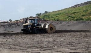 A bulldozer with a roller attachment is safeguarding the construction site's environmental health by efficiently flattening sandy terrain. In the background, a grassy hill lined with orange flags stretches under a cloudy sky, capturing the serene essence of an HEIC snapshot.