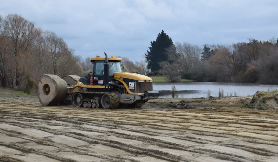 A yellow construction vehicle, equipped with a roller attachment, is leveling the ground near a small lake. In this area of sustainable forestry, fresh tracks are evident as trees with sparse foliage surround the scene under a cloudy sky.