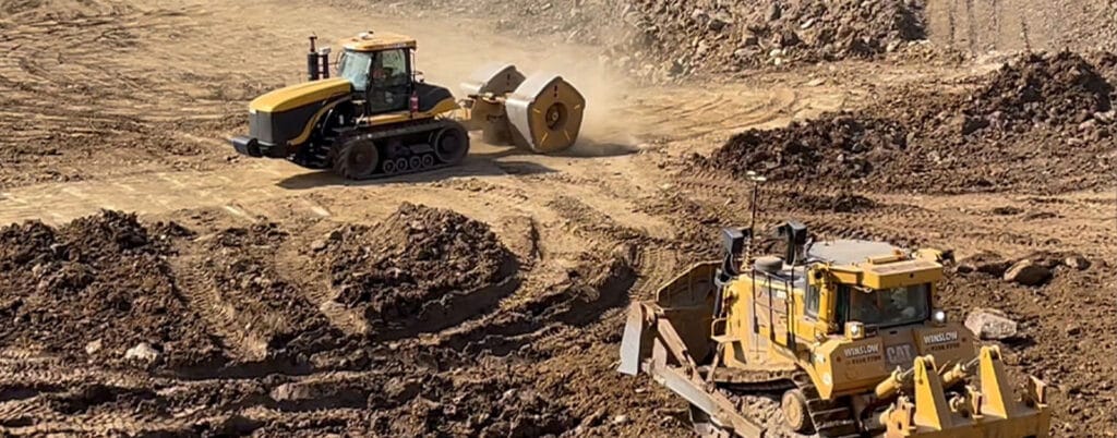 Two heavy machinery vehicles, a tractor and a bulldozer, work tirelessly on a large, dusty construction site. In the midst of developing power infrastructure, they move earth and shape the terrain with precision, surrounded by piles of dirt and uneven ground.