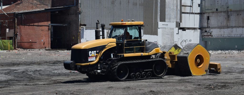 A large yellow and black construction vehicle with treaded tracks and a heavy roller attachment moves across a dirt surface in an industrial sector. Brick and concrete buildings stand in the background, creating a bustling business atmosphere.