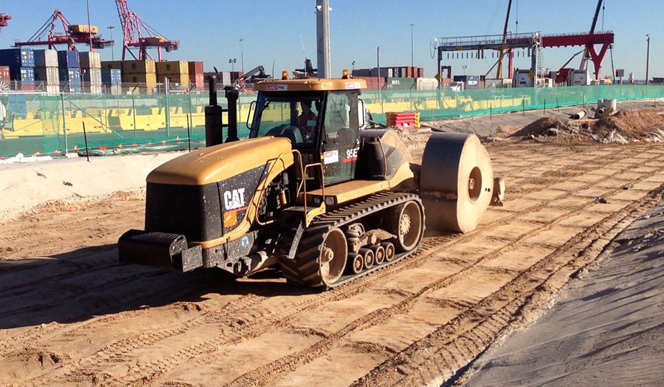 A bustling construction site showcases a Caterpillar vehicle with a large cylindrical attachment, efficiently flattening the ground. In this dynamic industrial sector scene, shipping containers and cranes line the background, all bordered by green fencing.