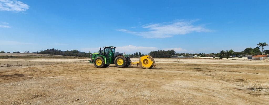 A green tractor with a yellow attachment sits on a vast, barren dirt field under a clear blue sky, symbolizing ongoing infrastructure development. In the background, trees and buildings stand as evidence of progress.