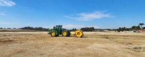 A green tractor with a yellow attachment sits on a vast, barren dirt field under a clear blue sky, symbolizing ongoing infrastructure development. In the background, trees and buildings stand as evidence of progress.