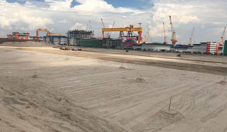 A large construction site with a flat sandy area in the foreground showcases numerous cranes reinforcing tomorrow's infrastructure. In the background, several multi-story buildings rise beneath a partly cloudy sky.