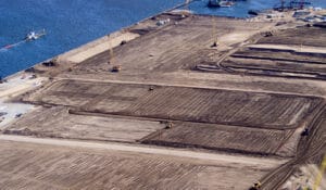 Aerial view of a large, sandy construction site advancing near a body of water. Several cranes are present along the shoreline, with machinery visible on the ground. The area is cleared and marked with parallel lines, indicating preparation for port development.