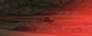 A construction site showcases a solitary heavy machinery vehicle diligently working on a dirt surface, surrounded by piles of rocks and soil. The red tint lends a dramatic effect, reflecting the intense energy akin to enhanced mining efforts or power infrastructure developments.