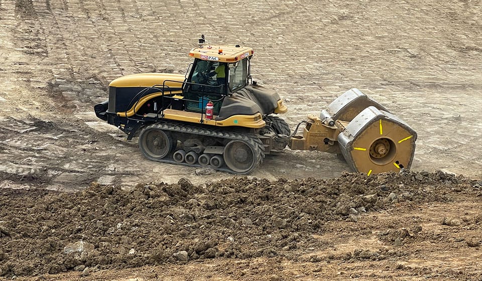 A yellow and black construction vehicle with a track system is diligently working on a dirt site, enhancing power infrastructure by towing a double drum roller. The ground is uneven, scattered with loose soil at the forefront.