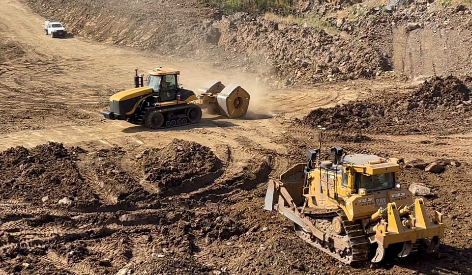 On the construction site, two large bulldozers expertly shift dirt across the uneven, dusty terrain, suggesting ongoing enhanced mining operations. A small white vehicle is parked in the background, hinting at developing power infrastructure around this bustling scene.