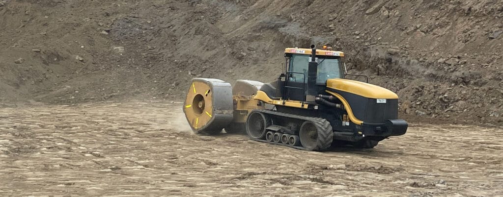 A large yellow and black construction vehicle with caterpillar tracks is flattening a dirt surface in a quarry, enhancing the mining infrastructure. Equipped with a heavy roller attachment, it works efficiently amid earth and rock walls surrounding the site.