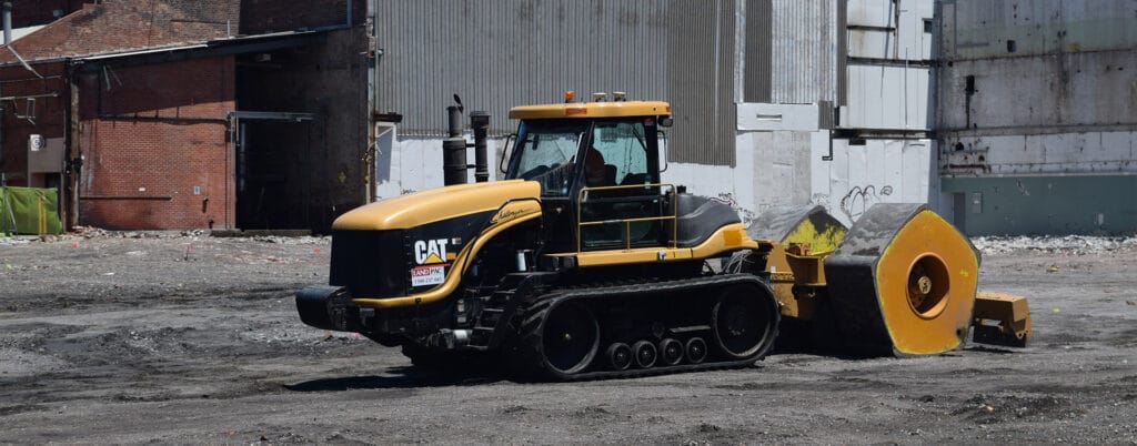 A yellow and black Caterpillar tractor, featuring intelligent compaction technology, pulls a heavy roller over a dirt-covered industrial site. In the background, brick and metal buildings rise above the ground cluttered with construction waste.